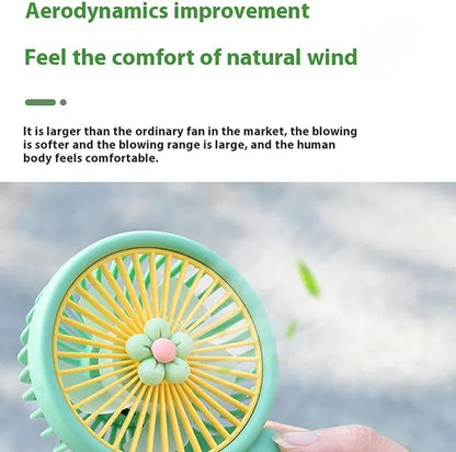 Handheld fan with green frame and yellow blades, held by a hand against a blurred outdoor background.